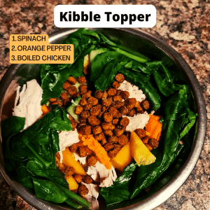 A bowl of kibble topped with spinach, boiled chicken and orange bell peppers.